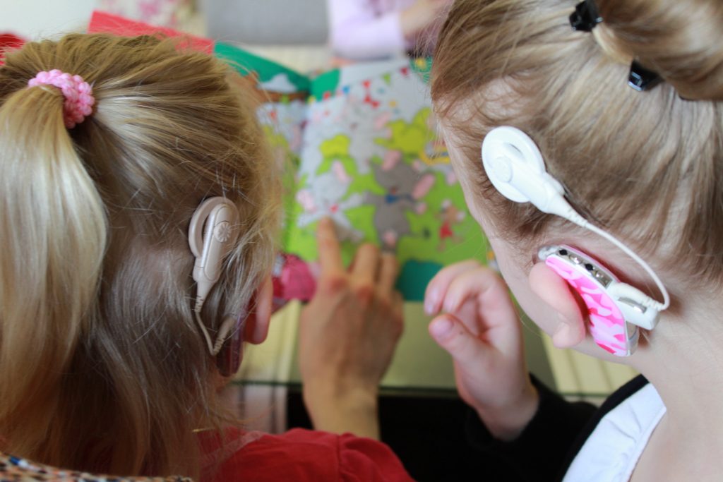 Lapsia takaapäin kuvattuna, molemmilla sisäkorvaistutteet. Lapset lukevat kirjaa.