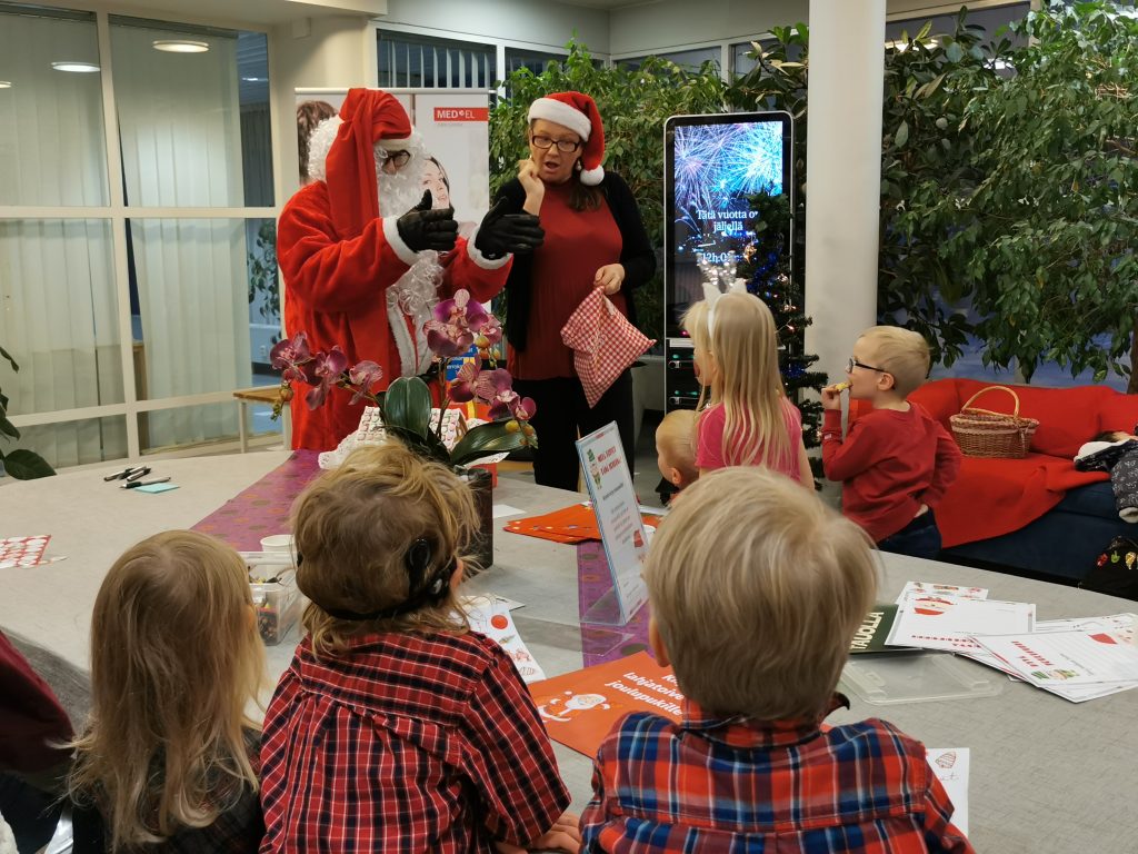 Joulupukki ja naisohjaaja viittovat lapsille, jotka ovat kerääntyneet askartelupöyvän ääreen. Yhdellä lapsista on sisäkorvaistute.