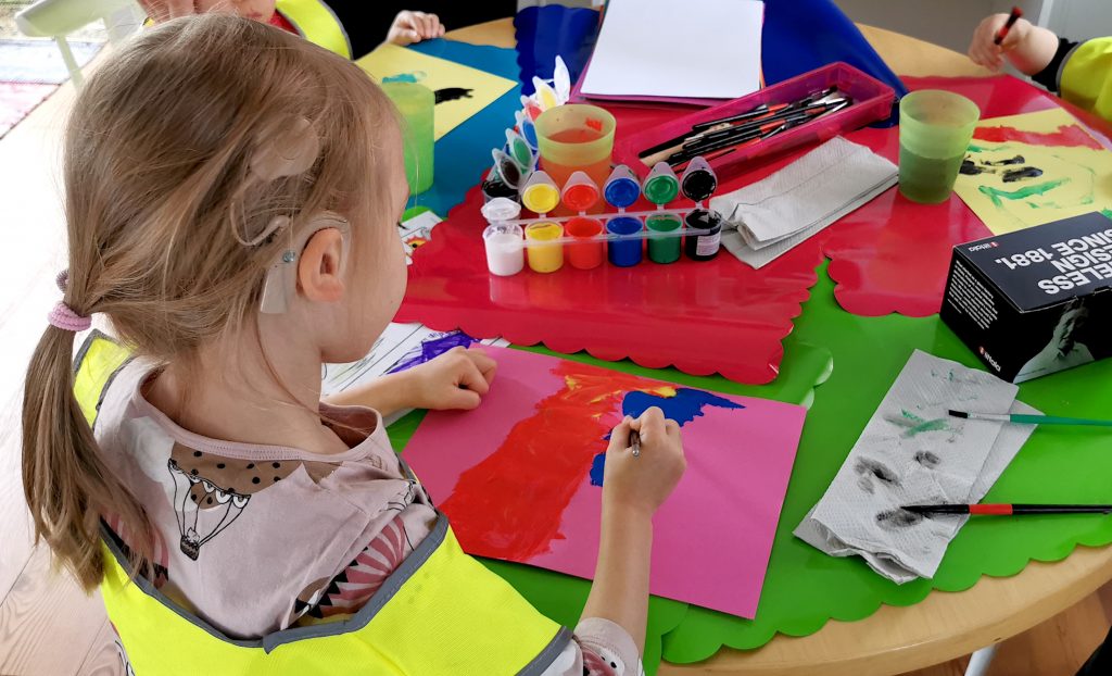 Kuvassa pieniä lapsia maalaamassa pöydän ääressä, etualalla olevalla lapsellä näkyy sisäkorvaistute.