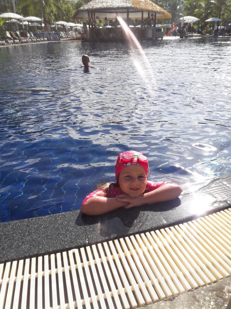 Kuvassa pieni tyttö uimalakki päässään nojaa uima-altaan reunaan, takana suihkuaa vettä