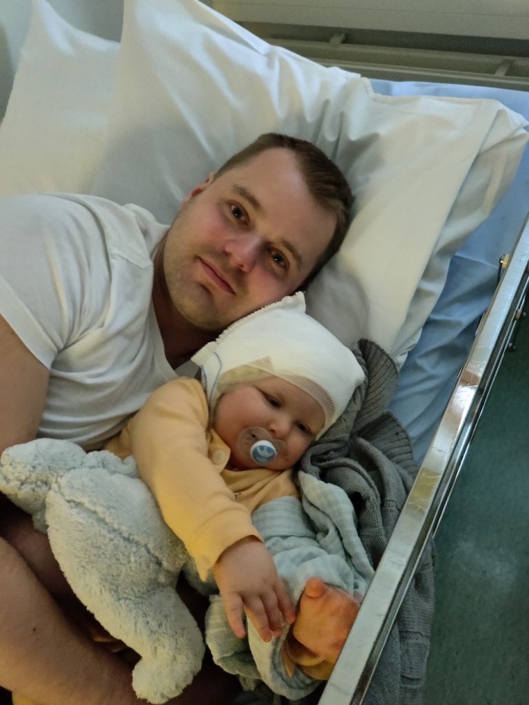 Isä makaa sairaalapedillä vauva sylissään, vauvalla pään ympärillä harso
