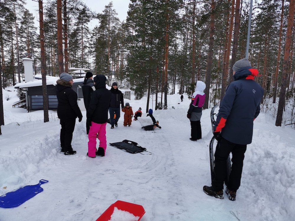 Kuvassa talvimaisema jossa joukko vanhempia on mäen päällä ja ympärillä pulkkia. Lapsia näkyy taaempana pulkkamäessä