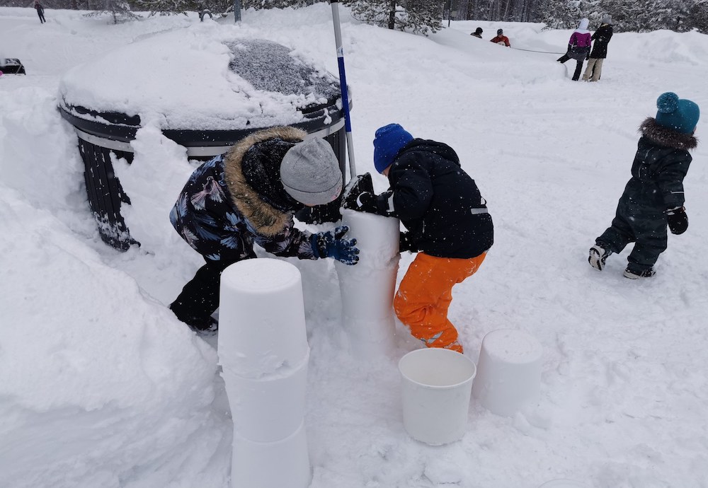 Kaksi lasta rakentaa lumesta ämpärin avulla pilaria. Taustalla näkyy muita lapsia ja perheitä