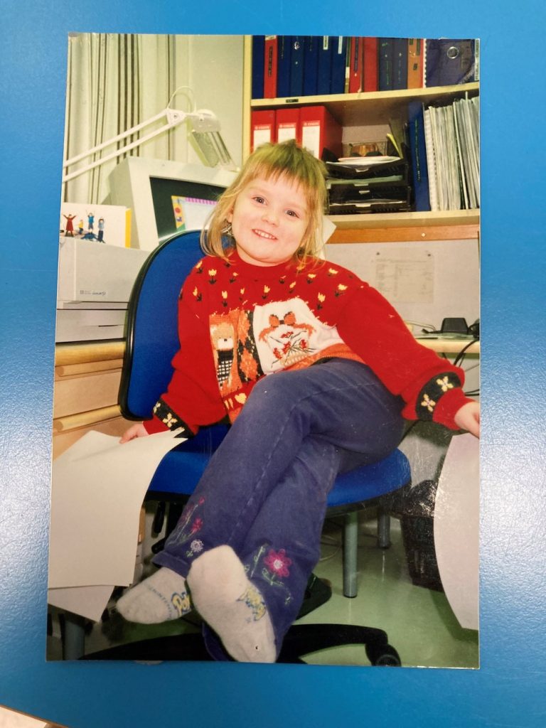Kuvassa pieni tyttö istuu hymyillen lääkärin tuolissa, takana tietokonepöytä