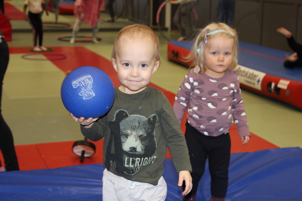 Kuvassa kaksi lasta liikuntasalissa, toisella lapsella kädessä pallo