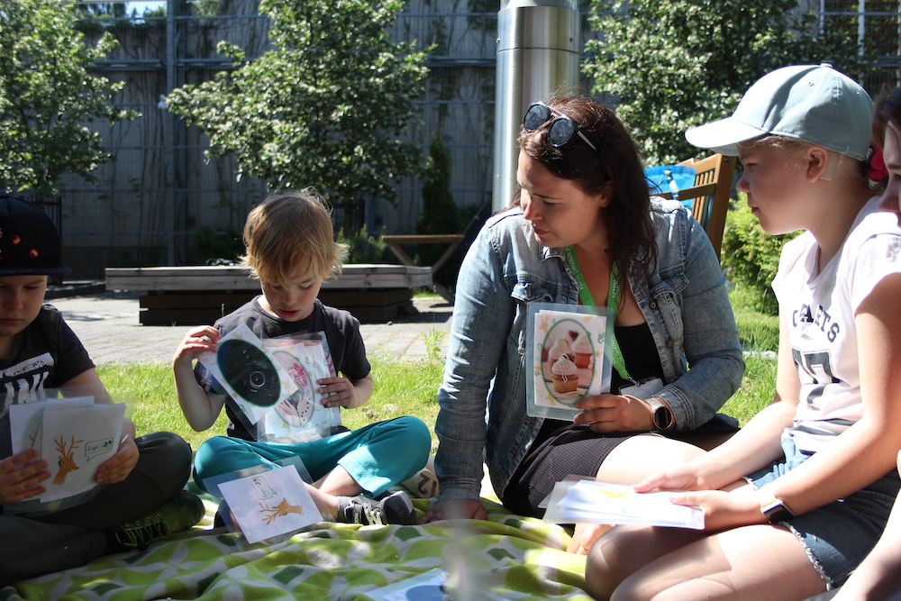 Kuvassa aikuinen ja lapsia istuu nurmella viltin päällä ja esittelee käsissään erilaisia kuvakortteja. Pienin poika on vuorossa, hän esittelee korttia, jossa näkyy kamera