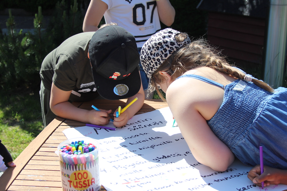 Kuvassa kaksi lasta nojaa pöytään ja kirjoittaa paperille väritusseilla ulkona puistossa.