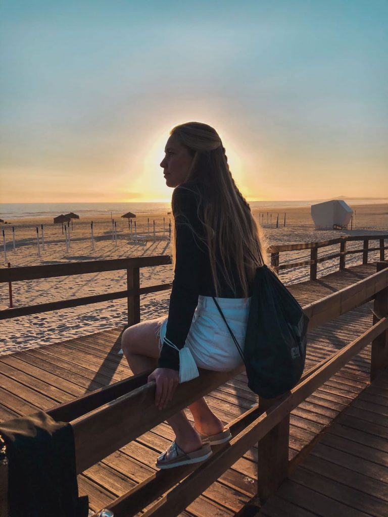 Kuvassa Sara-Elise istuu rannalla aidan päällä, aurinko paistaa takaapäin