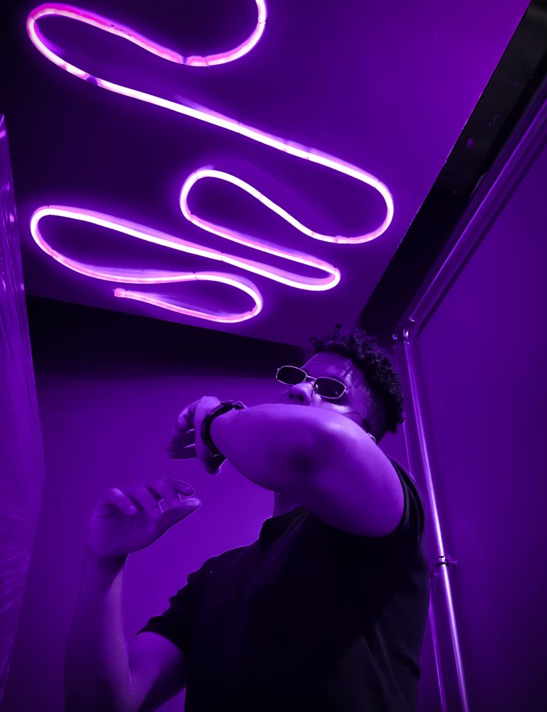 Kuvassa Juha-Miguel pimeässä, violetin valon täyttämässä huoneessa. Juha-Miguel näyttää tanssivan.