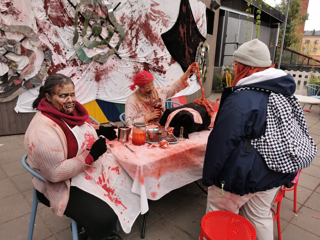 Kuvassa kaksi zombiksi pukeutunutta naista "verisen" pöydän ääressä, edessä heitä katselee nuori osallistuja. Zombien edessä pöydällä on verisiä purkkeja ja nukke