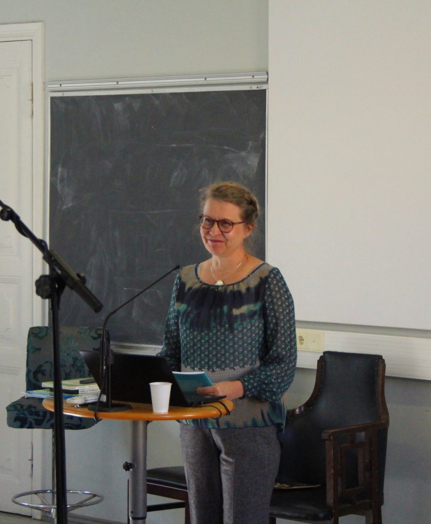 Kuvassa Leena Hasselman seisoo auditoriossa puhujan paikalla edessä.