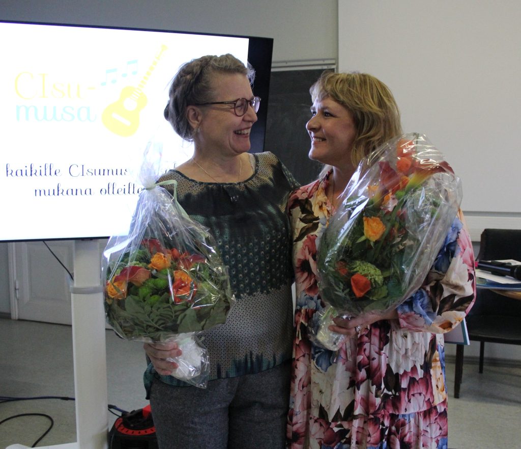 Kuvassa kaksi henkilöä (Leena Hasselman ja Janet Grundström) seisovat vierekkäin ja katsovat toisiinsa. Kädessään heillä on kukkakimput.