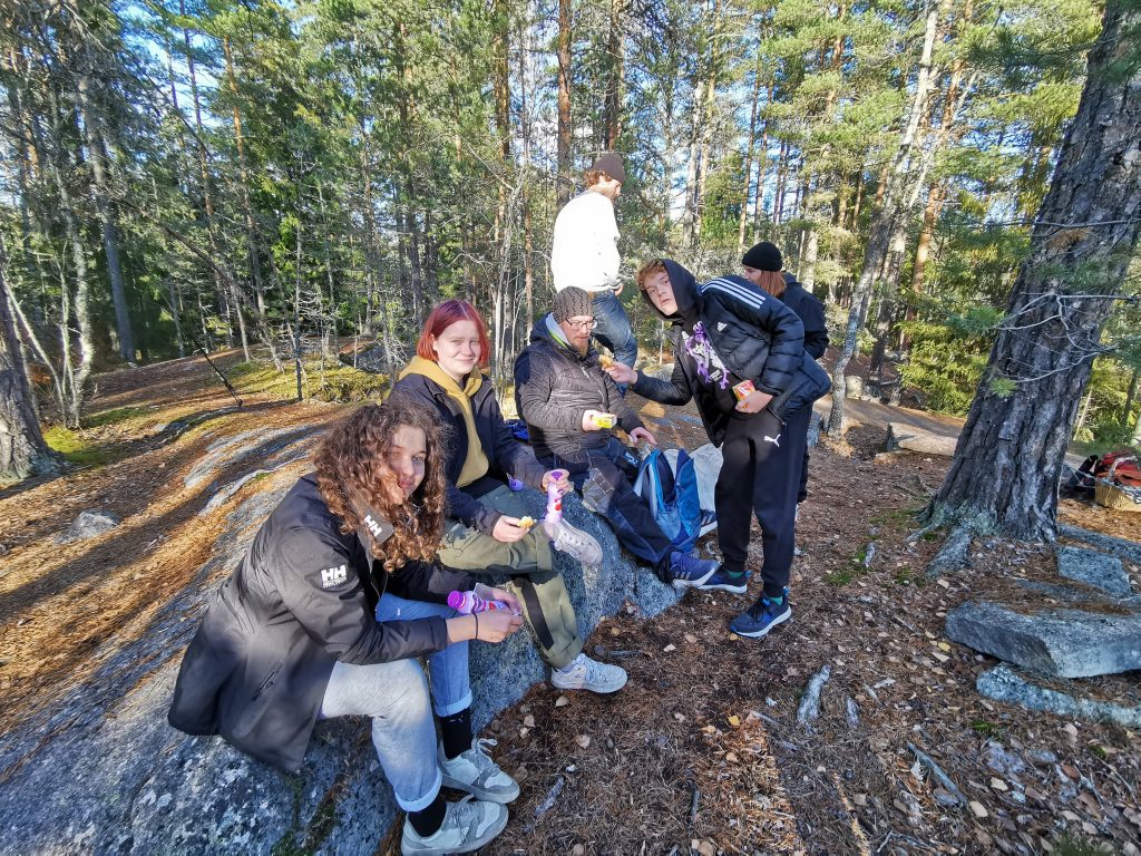 Kuvassa kolme nuorta ja yksi ohjaaja poseeraavat kameralle hymyillen, he istuvat ison kiven päällä keskellä metsää. Takana näkyy muista osallistujia