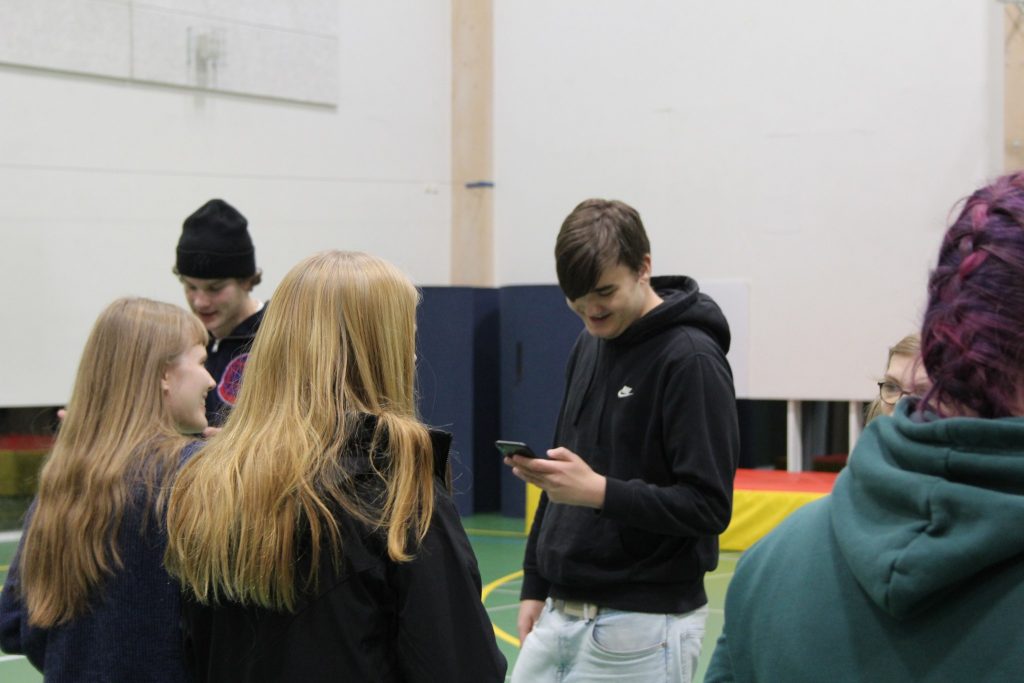 Kuvassa nuoria seisomassa vastakkain toisiaan, keskellä kuvaa oleva teinipoika katsoo hymyillen kädessään olevaa kännykkää
