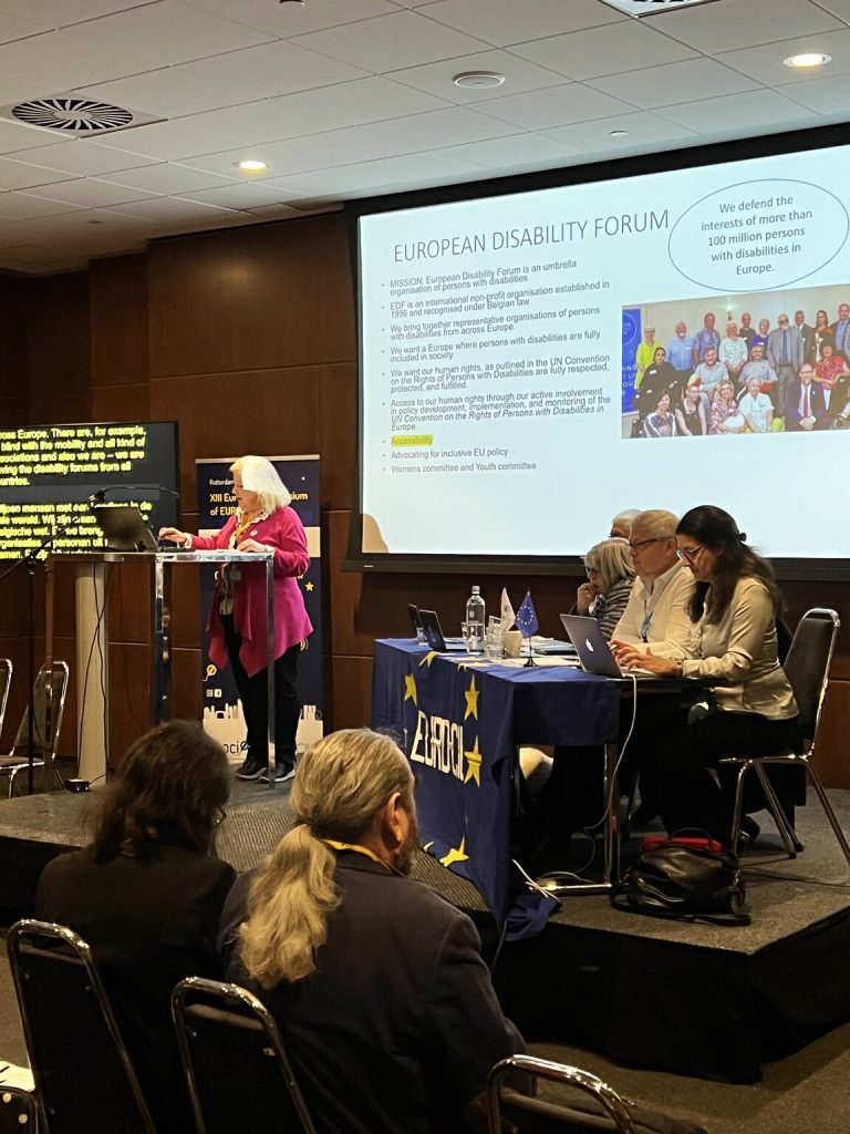Kuvassa puhuja korokkeella, hänen vieressään pöytä jonka ääressä on Euro-ciun edustajia. Takana näkyy esitys kankaalla, esityksessä kerrotaan European Disability Forumista.