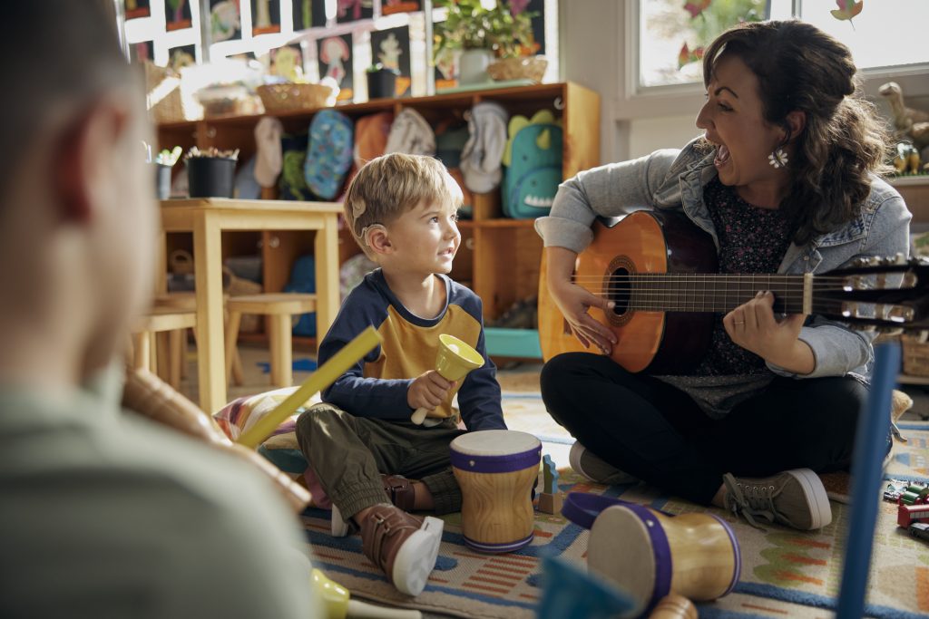 Kuvassa pieni poika päässään Nucleus 8 -puheprosessorit istuu leikkihuoneen lattialla ja katsoo vieressään laulavaa ja kitaralla soittavaa naista. Lapsen edessä rumpuja ja muita soittimia