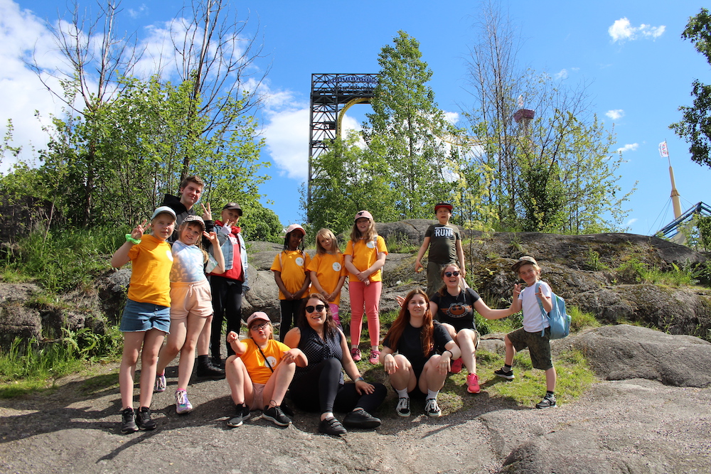 Musaleirin osallistujat ja ohjaajat poseeraavat Linnanmäen huvipuiston edessä kalliolla. Osalla lapsista on päällään oranssi paita.