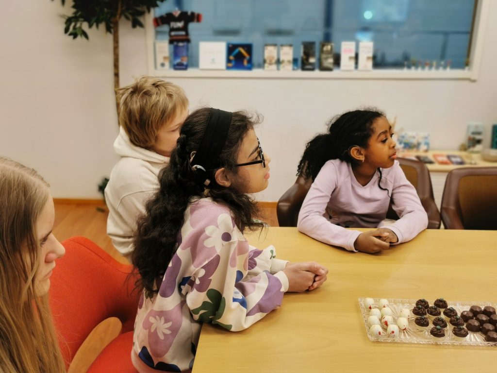 Kuvassa lapsia istuu pöydän ääressä ja katsoo oikealle. Lapsista parilla näkyy sisäkorvaistutteet. Pöydällä on suklaata.
