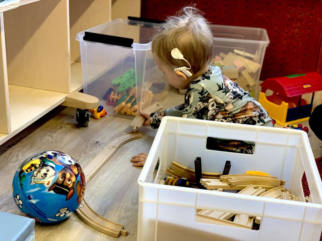 kuvassa lapsi leikkimässä kahden lelulaatikon välissä, lapsen päässä on valkoinen sisäkorvaistute