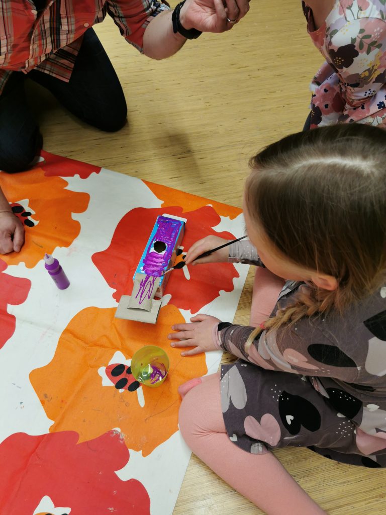 Pieni tyttö istuu lattialla ja maalaa maitopurkista tuunattua soitinta violetiksi.
