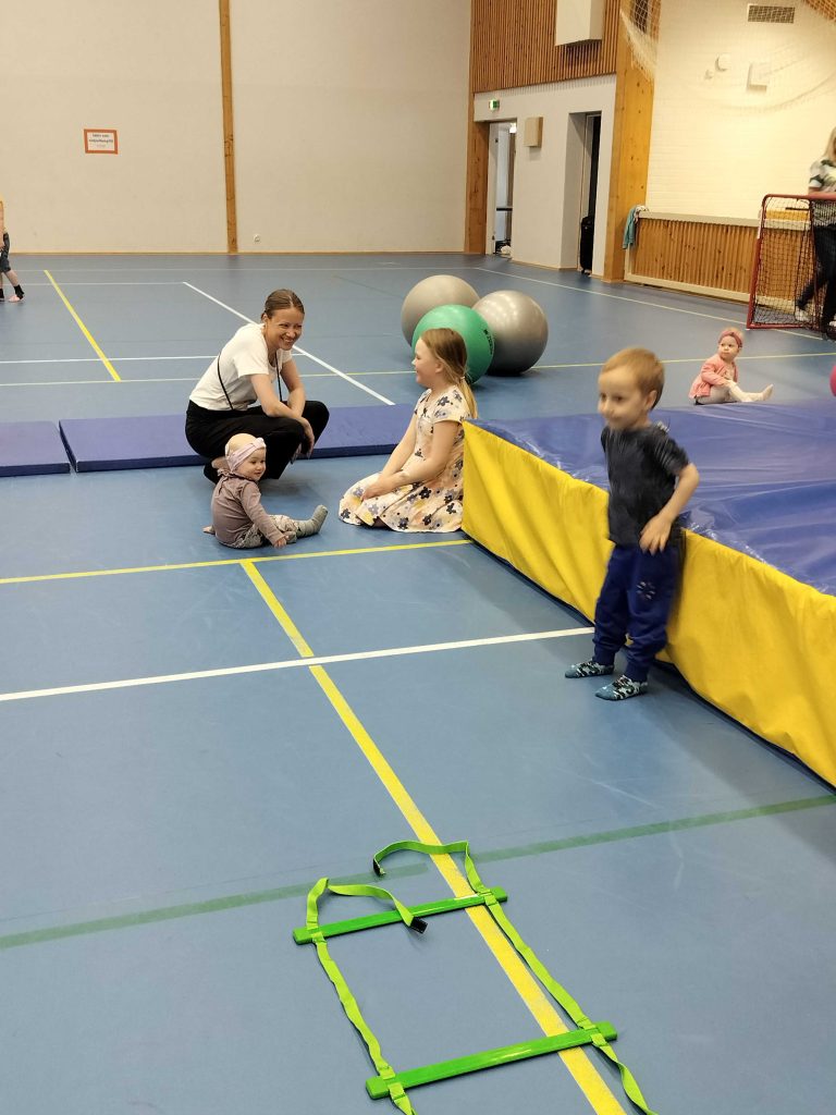 Kuvassa on iso liikunasali, jossa on jumppapatja ympärillä lattialla lapsia ja yksi aikuinen kyykyssä.