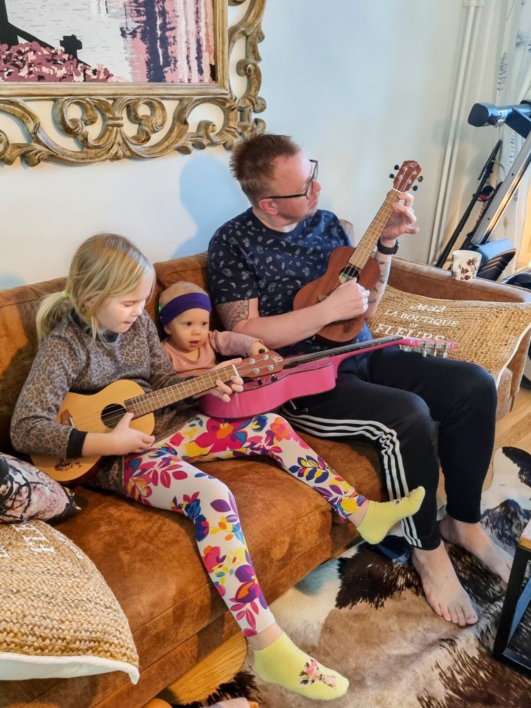 Kuvassa ruskealla sohvalla istuu isä ja kaksi tytärtä. Heillä kaikilla on ukulele kädessään.