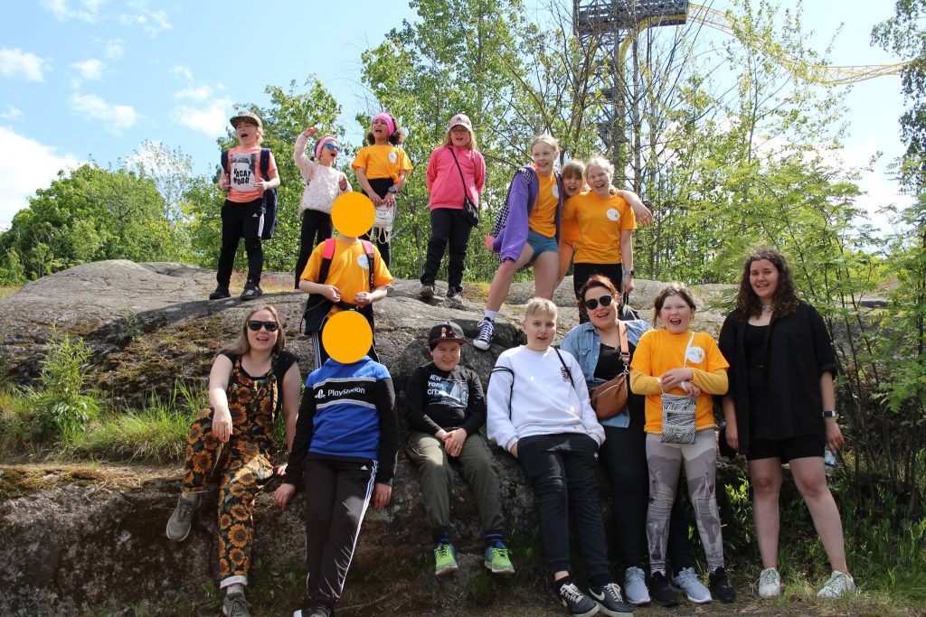 Joukko musaleiriläisiä ja kolme ohjaajaa poseeraavat ryhmäkuvassa Linnanmäen edessä kalliolla. Lähes kaikilla on suu auki ja hymyssä