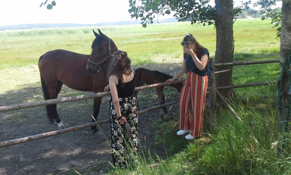 Mirella ja toinen nuori nainen selin kameraan päin, he seisovat hevosten aitauksen vieressä. Toinen naisista peittää kasvojaan kädellä.