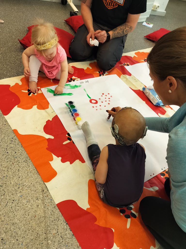 Kaksi pientä lasta maalaavat sormimaaleilla isolle valkoiselle paperille ja heidän vieressään istuu aikuiset auttamassa. Kuva: CIsumusa-viikonloppu syksy 2023.