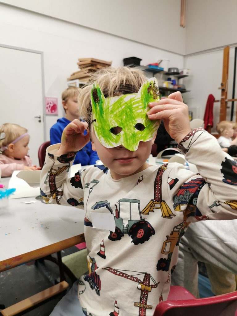 Pieni poika katsoo kameraan kasvoillaan vihreä itsetehty naamio. Takana näkyy muita lapsia pöydän ääressä.