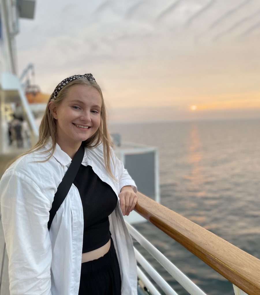 Jenna poseeraa hymyillen laivan kaidetta vasten, taustalla näkyy auringonnousu horisontissa.