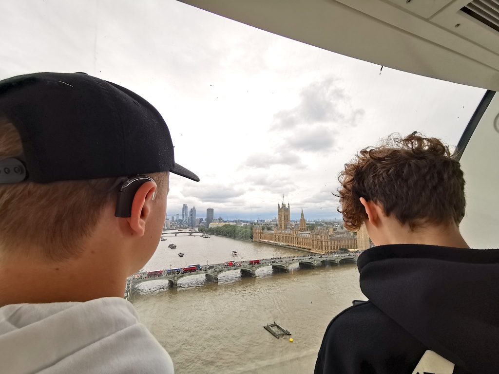 Kaksi nuorta poikaa, joilla on sisäkorvaistutteet, kuvattuina takaapäin. Pojat katsovat ikkunasta ulos, ikkunasta näkyy Big Ben -kellotorni, Thames-joki ja pilvinen taivas.
