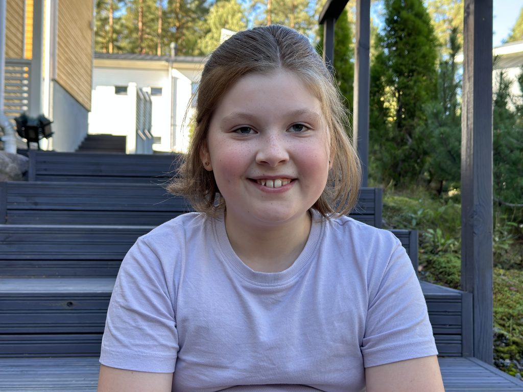 Olivia istuu talon portailla ja hymyilee lähikuvassa kameralle.