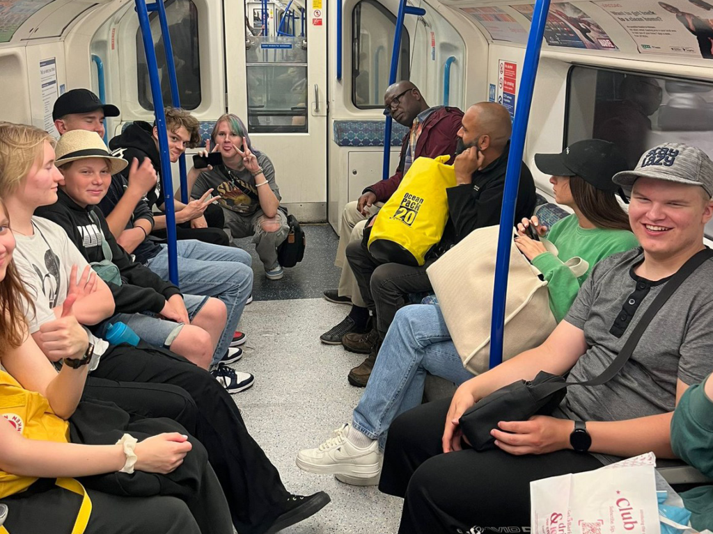 Nuoria istuu Lontoon metrossa, osa hymyilee ja poseeraa kameralle. Muut matkustajat katsovat kamerasta poispäin. 