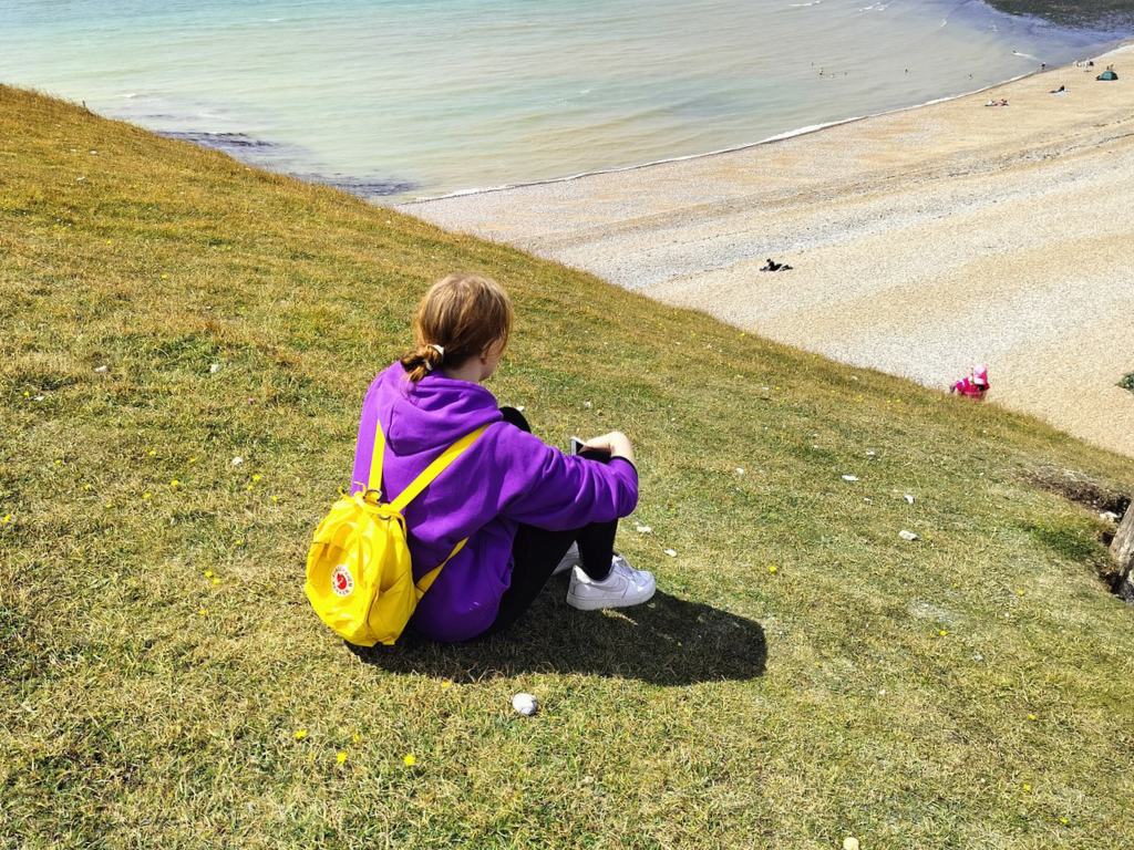 Teinityttö istuu rinteellä ja katsoo rannalle päin. Tyttö on selin kameraan päin.