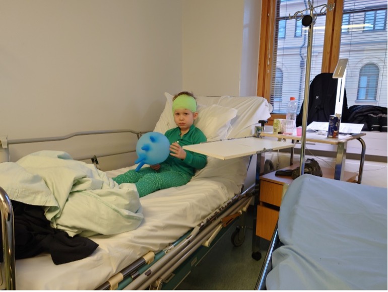 Lapsi istuu sairaalavuoteella vihreässä sairaala-asussa sininen ilmapallo sylissään. Päässä lapsella, on side aivokalvotulehhduksen jälkeisen sisäkorvaistuteleikkauksen jälkeen.