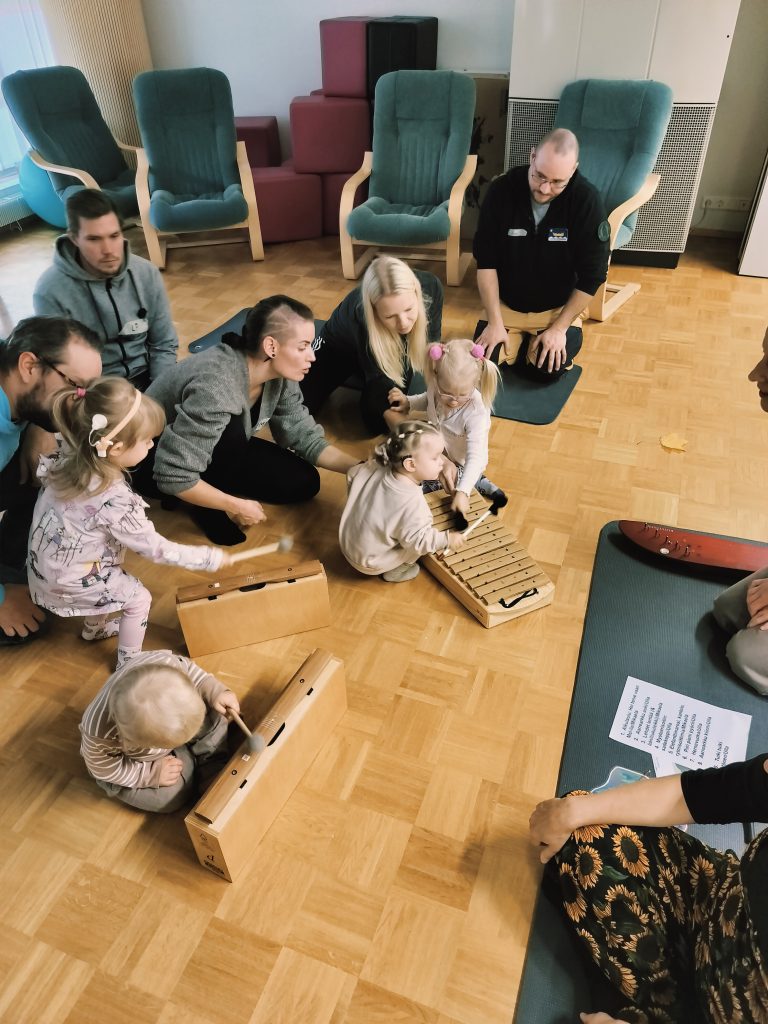 Neljä lasta ja neljä aikuista soittamassa lyömäsoittimia lattialla