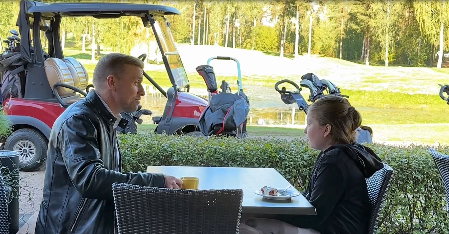 Isä ja tytär istuvat pöydän ääressä ulkoterassilla, isällä kahvikuppi ja lapsella edessä kakkulautanen. Taustalla golf-kärryjä ja nurmikkoa.