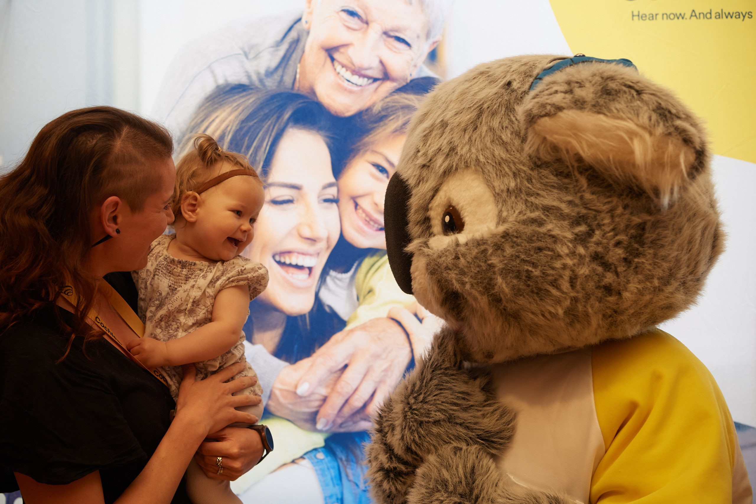 Lapsi on äidin sylissä ja hymyilee, katsoo edessään olevaa koala-asuun pukeutunutta maskottia.