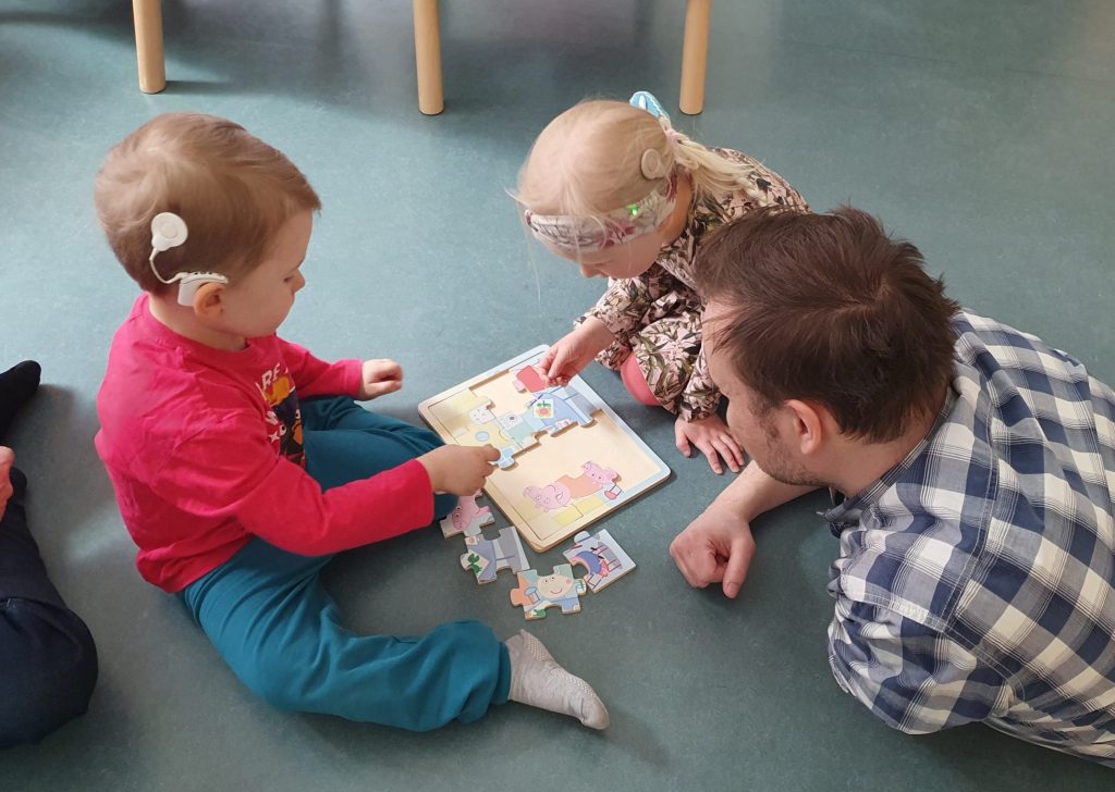 Kaksi pientä lasta leikkii palapelillä aikuisen kanssa lattialla. Lapsilla on SI:t.