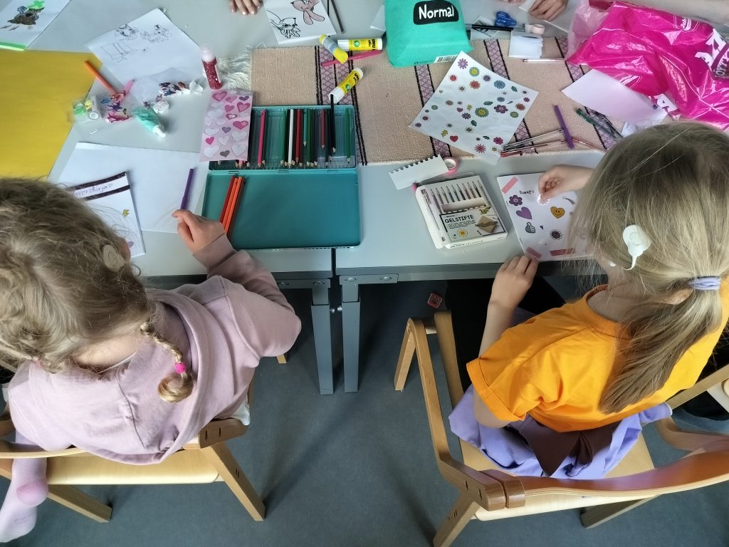 Ylhäältäpäin otetussa kuvassa kaksi SI:ta käyttävää lasta lisää vihkoihinsa erilaisia värikkäitä tarroja. Pöydällä lojuu kyniä, tarroja, liimoja ja muita askartelutarvikkeita.
