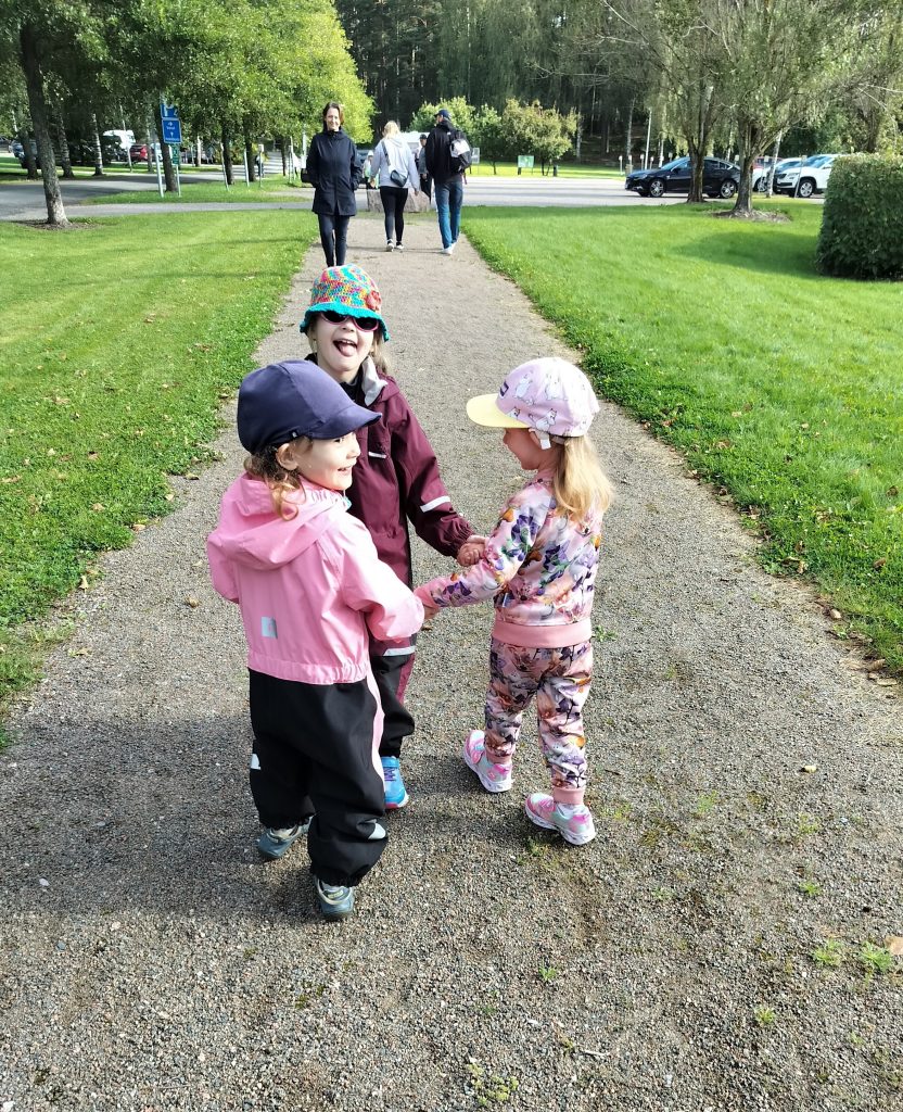 Etualalla kolme pientä lasta pitelee toisiaan käsistä, hymyilee ja nauraa. Taustalla näkyy vanhempia, yksi heistä kääntynyt lasten suuntaan.