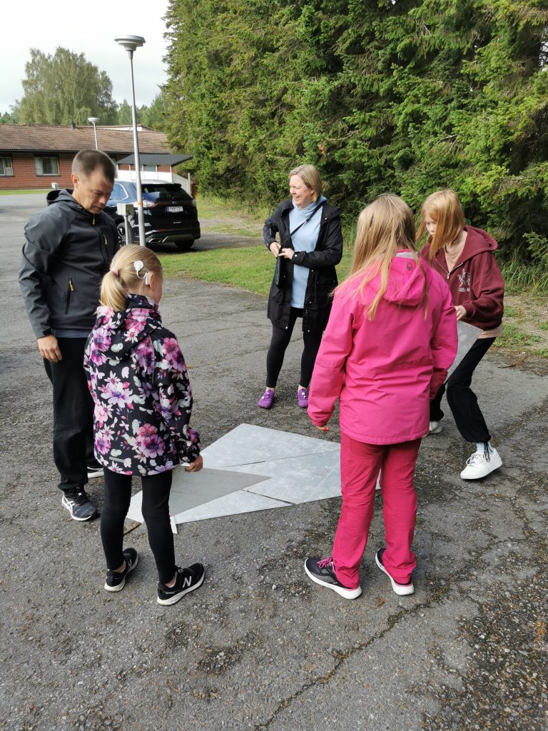 Perhe seisoo piirissä eri muotoisten laattojen ympärillä ja hymyilee. 