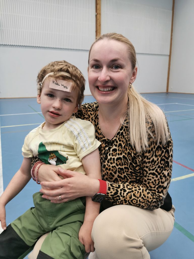 Äiti ja lapsi lähikuvassa, he hymyilevät. Lapsella otsallaan lappu, jossa lukee "pikku Myy".