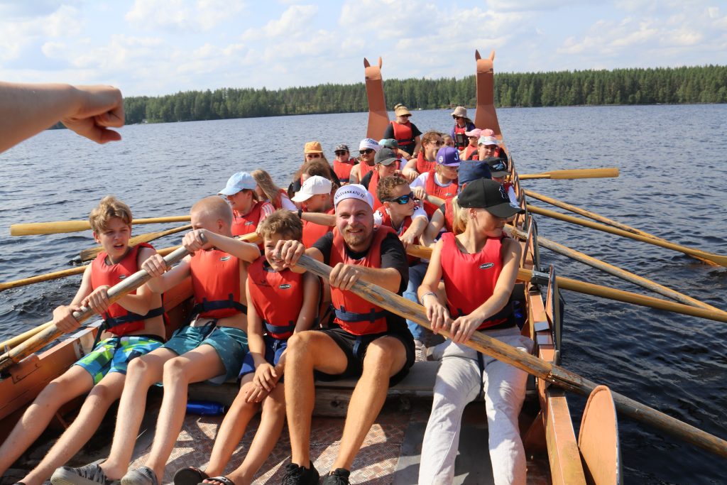 Nuoria ja ohjaajia ison viikinkiveneen kyydissä järvellä, kaikilla airot kourassa. 