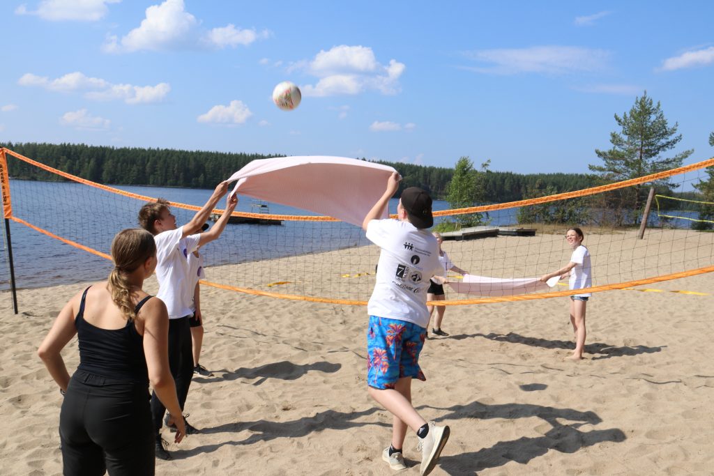 viisi ihmistä pelaa beachvolleytä pyyhettä käyttäen. 