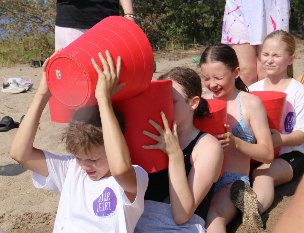 Nuoria istuu jonossa hiekalla isot ämpärit käsissään. Nuoret siirtävät vettä ämpäristä toiseen päänsä yli.