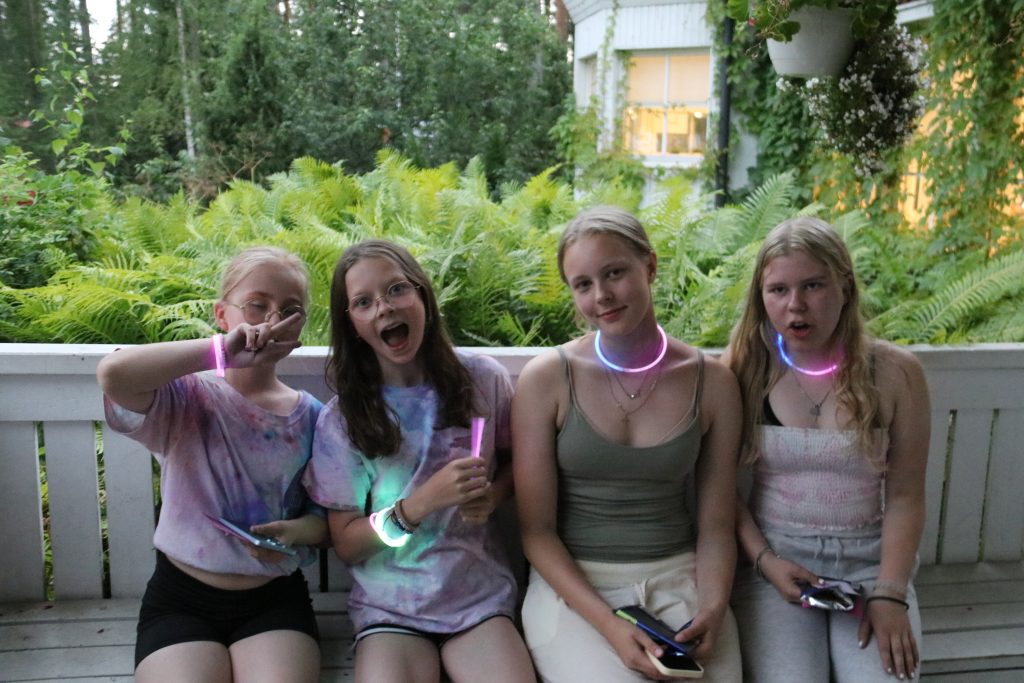 Neljä nuorta istuu vierekkäin kuistilla ja taustalla näkyy valkoinen kaide ja saniaisia.