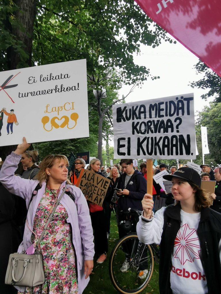LapCI ry:n toiminnanjohtaja ja järjestöassistentti seisomassa mielensosoituksessa, molemmilla tympiintyneet ilmeet. Heidän pitämissään kylteissä lukee "Ei leikata turvaverkkoa!" ja "Kuka meidät korvaa? EI kukaan."