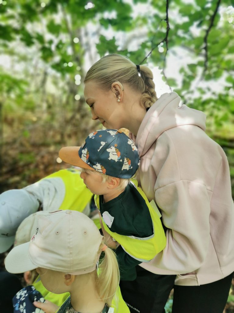 Ohjaajanuori pitää pientä osallistujaa sylissään ja hymyilee. Ympärillä muita lapsia, taustalla näkyy metsää.